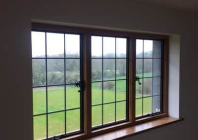 Three‑pane wooden‑framed heritage metal‑style window with black grid muntins, overlooking a green countryside landscape.
