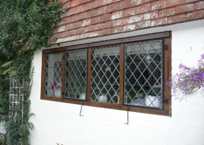 White‑brick cottage wall with a dark timber‑framed diamond‑paned heritage metal‑style window, surrounded by climbing greenery and a hanging flower basket.