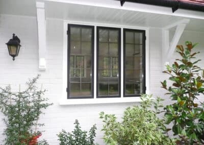 Black metal‑framed heritage window with grid glazing set into a white brick exterior with garden shrubs.