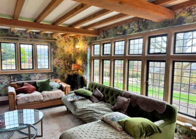 Cozy living room with large multi‑pane windows, exposed beams and nature‑themed décor overlooking a garden.