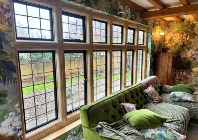 Indoor sitting area with light‑wood framed multi‑pane windows, forest wallpaper and green velvet seating facing a garden view.